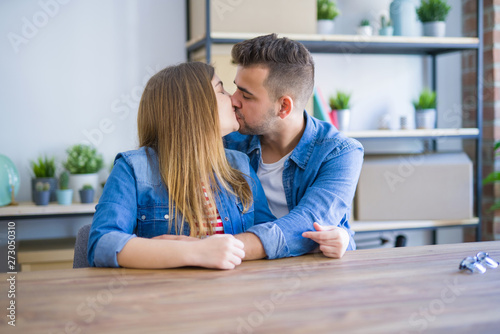 Young beautiful couple sitting on the table at home, hugging in love very happy for moving to new home with cardboard boxes behind them