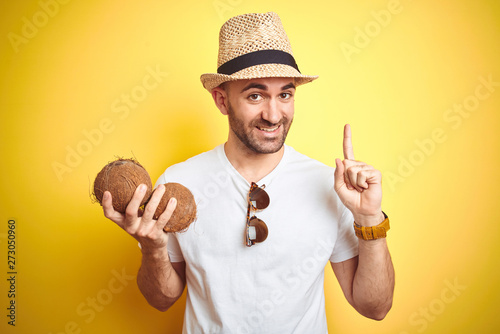 Young man wearing summer hat and holding coconut fruit over yellow background surprised with an idea or question pointing finger with happy face, number one