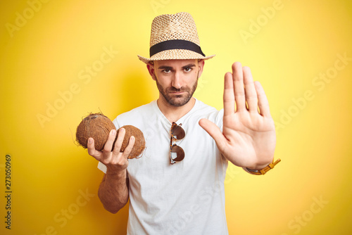 Young man wearing summer hat and holding coconut fruit over yellow background with open hand doing stop sign with serious and confident expression, defense gesture