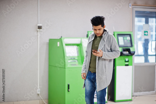 Young stylish asian man with mobile phone against row of green ATM.