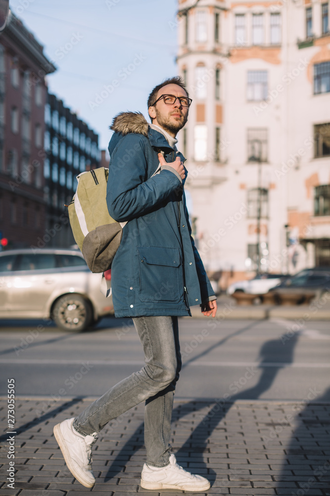 Vertical photo of handsome adult male in blue jacket and white sneakers. Pretty man with beard and glasses walking on the street and right hand holding green backpack. Lifestyle in big city. 