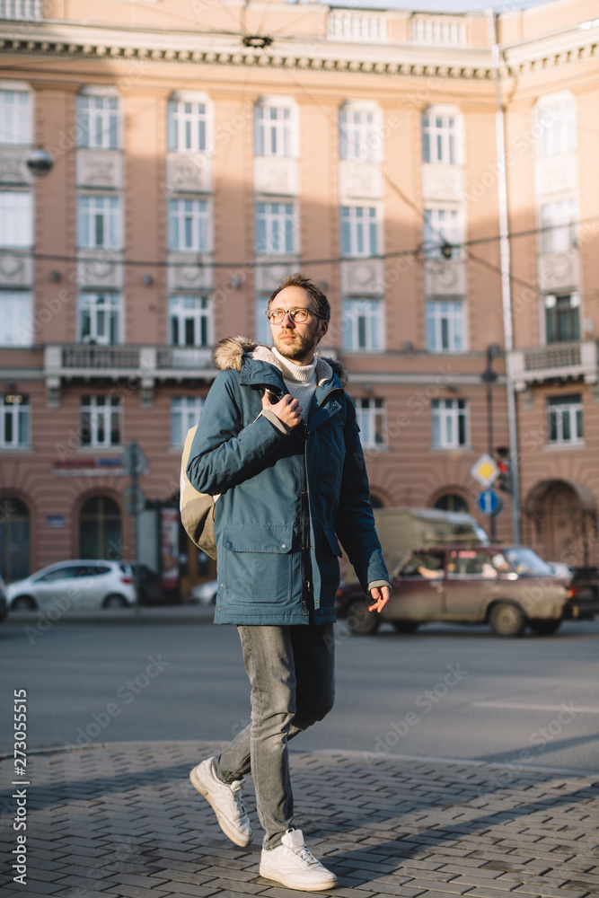 Photo of attractive adult male in blue jacket and white sneakers. Pleasant man with beard and glasses goes to work and right hand holding green backpack. Hipster in urban environment. Winter season.