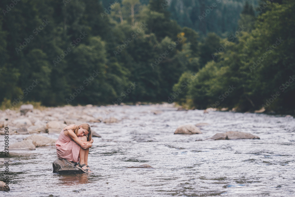 Fototapeta premium Young lonely girl sitting at stone in the river among mountains in summer.