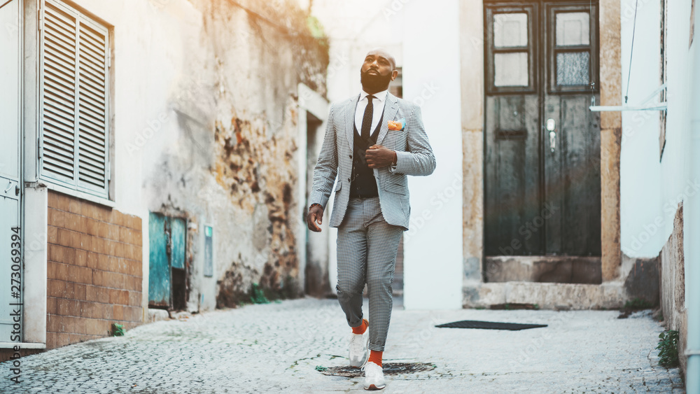 An elegant bald bearded African man in a fashion costume with tie and orange socks is walking down the antique street of a European city; a fancy black guy in a suit is leaving the door of his house An elegant bald bearded African man in a fashion costume with tie and orange socks is walking down the antique street of a European city; a fancy black guy in a suit is leaving the door of his house