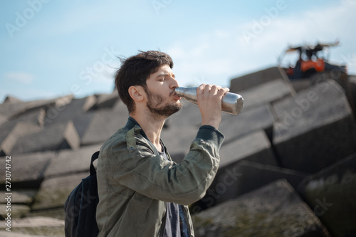 young man with aluminium thermo bottle on the beach on the background of big square stones 