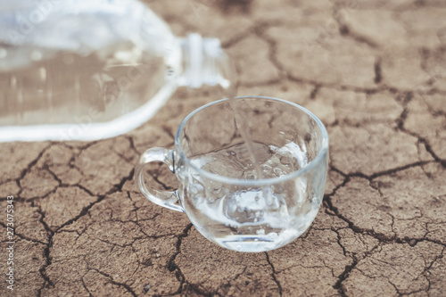 Concept of thirst, dehydration, lack of water. Water pouring into a glass cup on the background of dried cracked ground soil.
