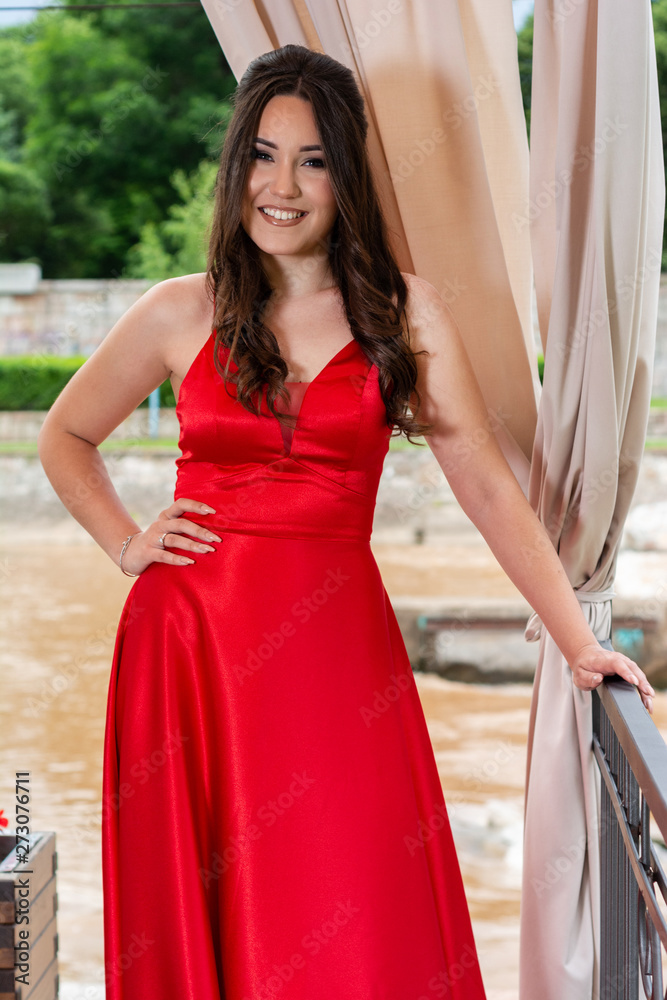 Young girl in red dress and teeth smile posing and holds a hand on hips in front of the iron fence and curtains in the garden on a summer day. Fashion and beauty concept. Close up, selective focus