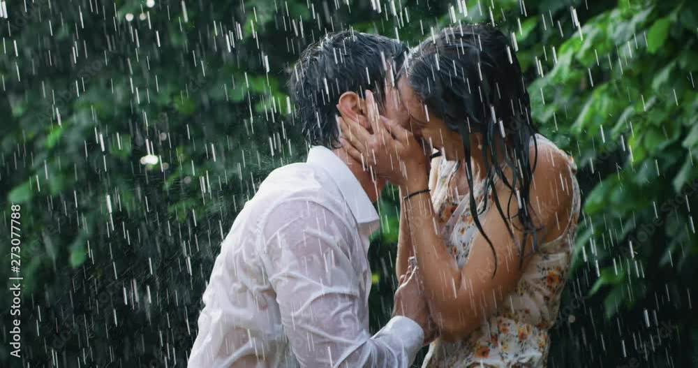 Slow motion of young carefree couple in love are hugging and kissing under the rain on a background of green trees.