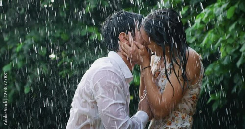 Slow motion of young carefree couple in love are hugging and kissing under the rain on a background of green trees.