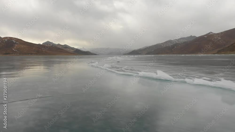 Low angle drone flight over broken ice at frozen Yamdrok lake with reflection of mountains and grey clouds, on high altitude plateau in Tibet Low angle drone flight over broken ice at frozen Yamdrok lake with reflection of mountains and grey clouds, on high altitude plateau in Tibet