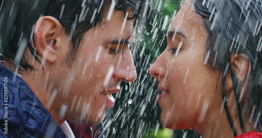 Slow motion of young carefree couple in love are wearing protection capes hugging and kissing under the rain on a background of green trees.