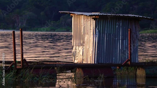 Small metal cabin stands next to fishing cages by the shore of Lake Victoria. Orange sky reflected on surface. Lush forest in background. Sunset in Kalangala, Uganda. photo