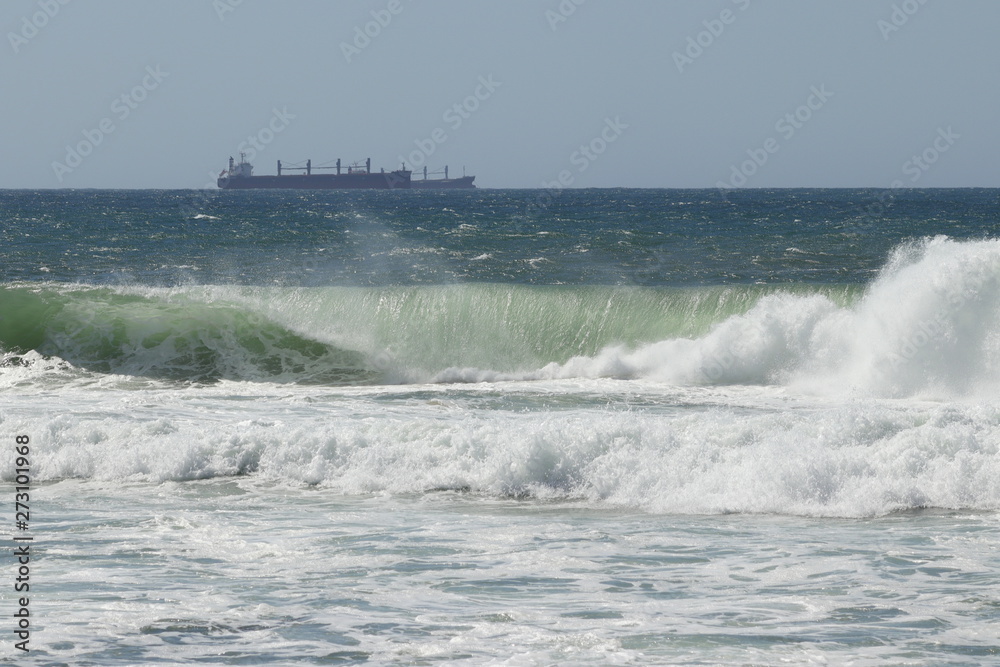 Big storm waves hitting the beach of the sea. Cargo ships at anchor on the background. Sunny day.