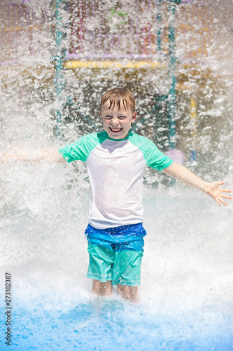 Young boy getting soaking wet while at an outdoor water park. Lots of water splashing water behind the boy. He is smiling and anticipating getting wet and drenched