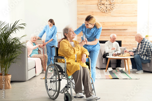 Nurse giving glass of water to elderly woman in wheelchair at retirement home. Assisting senior people photo