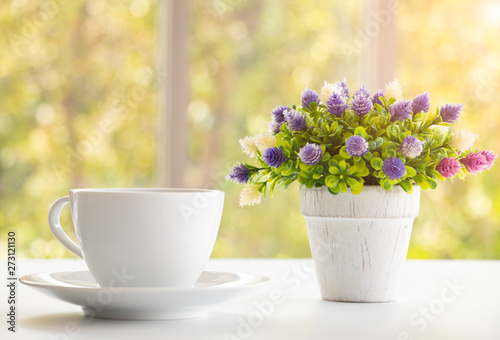 A cup of coffee on white office desk with lovely vase of small flower with morning light flare. A good starting in the early morning to get ready for a day