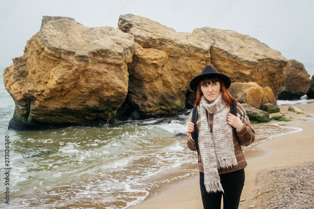 Portrait of a young red-haired woman in a hat and a scarf with a backpack against the background of the rocks against the beautiful sea, on the coast, on the horizon. Tourism, rest, lifestyle.