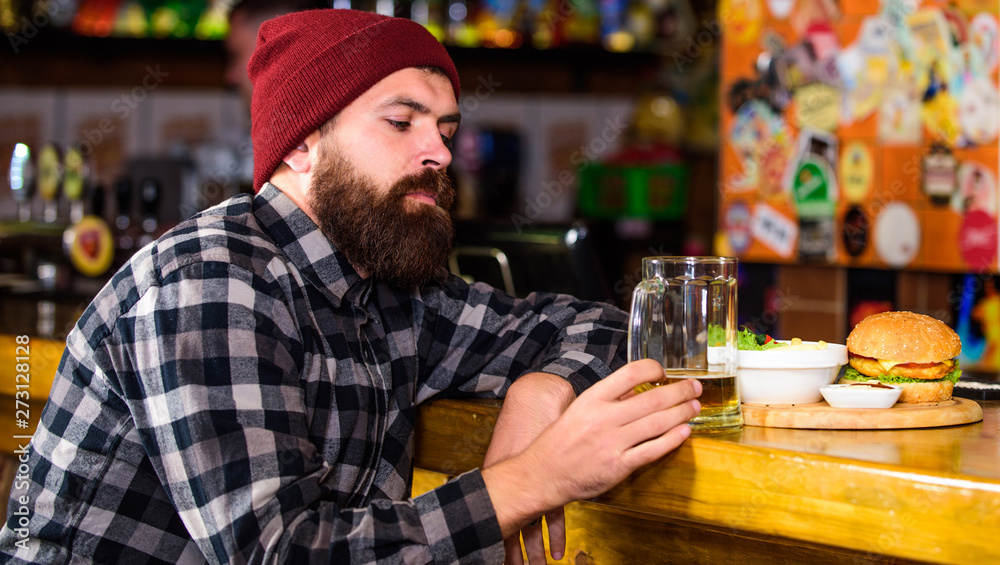 Brutal hipster bearded man sit at bar counter. Hipster relaxing at pub. Pub is relaxing place to have drink and relax. Man with beard drink beer eat burger menu. Enjoy meal in pub. High calorie snack