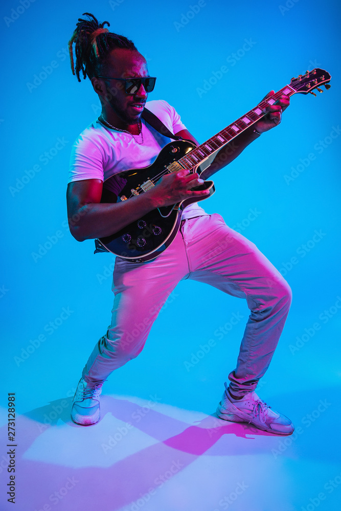 Fototapeta premium Young african-american musician playing the guitar like a rockstar on blue studio background in neon light. Concept of music, hobby. Joyful attractive guy improvising. Retro colorful portrait.