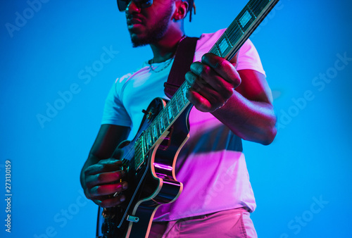 Young african-american musician playing the guitar like a rockstar on blue studio background in neon light. Concept of music, hobby. Joyful attractive guy improvising. Retro colorful portrait. photo