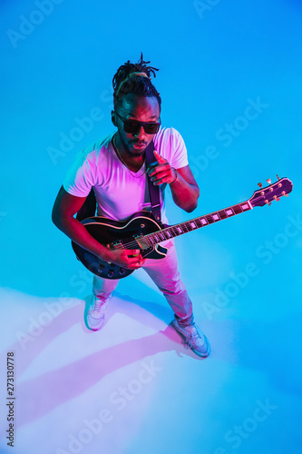 Young african-american musician playing the guitar like a rockstar on blue studio background in neon light. Concept of music, hobby. Joyful attractive guy improvising. Retro colorful portrait. © master1305