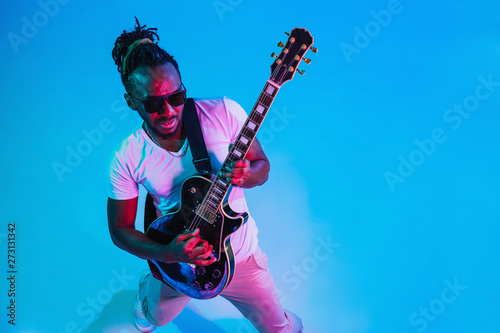 Young african-american musician playing the guitar like a rockstar on blue studio background in neon light. Concept of music, hobby. Joyful attractive guy improvising. Retro colorful portrait.