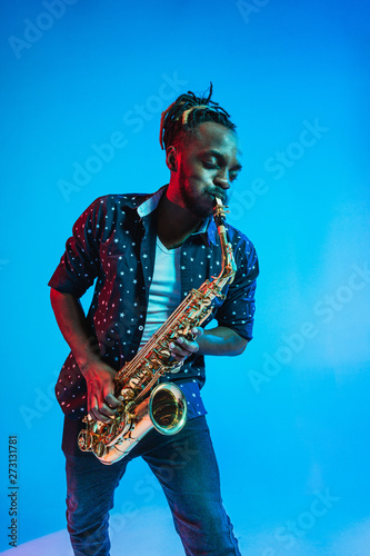 Photography Young african-american jazz musician playing the saxophone on blue studio background in trendy neon light