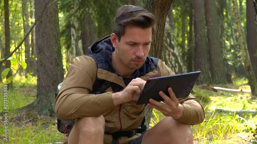A young handsome hiker sits on the ground in a forest and works on a tablet A young handsome hiker sits on the ground in a forest and works on a tablet