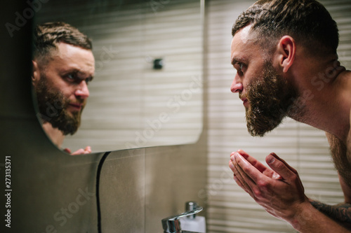 Fotografie Young bearded hipster man washing his face with water in the bathroom while looking himself in the mirror