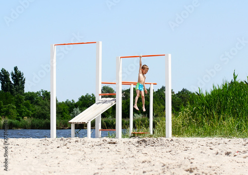 Little boy is hanging on one hand on sport equipment on the beach after pulling up