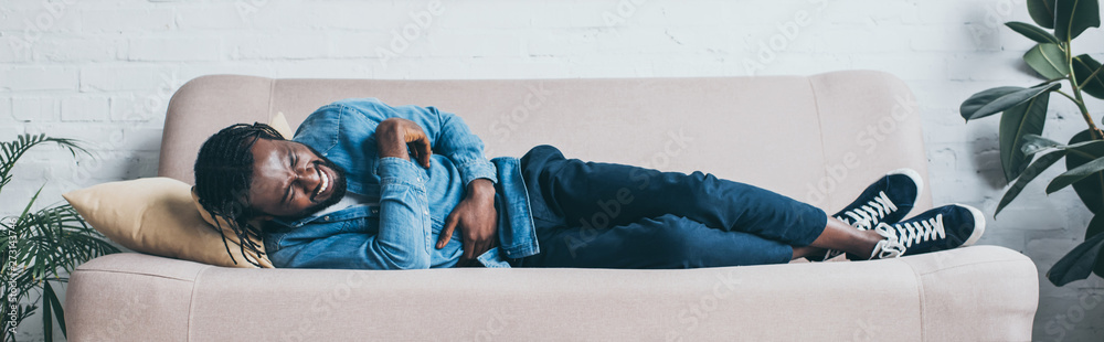panoramic shot of african american man suffering from abdominal pain while lying on sofa panoramic shot of african american man suffering from abdominal pain while lying on sofa