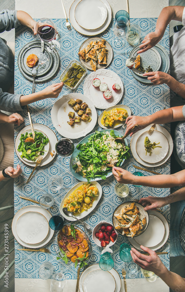 Mediterranean style dinner. Flat-lay of table with salad, starters, pastries over blue table cloth with hands holding drinks, sharing food, top view. Holiday vegetarian party concept