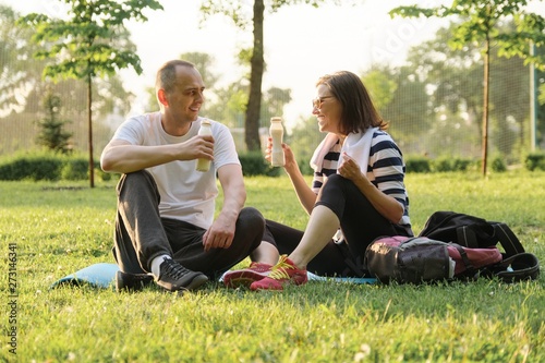 Happy mature couple sitting in the park on fitness mat, resting drinking yogurt after sports exercises.