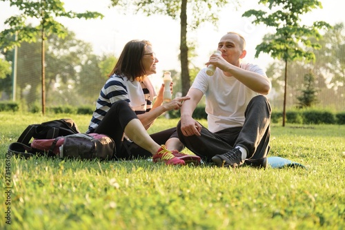 Happy mature couple sitting in the park on fitness mat, resting drinking yogurt after sports exercises.