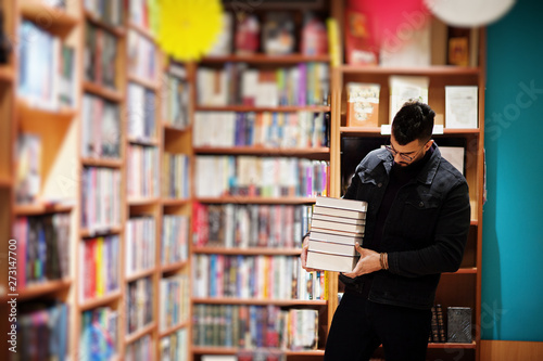 Tall smart arab student man, wear on black jeans jacket and eyeglasses, at library with stack of books.