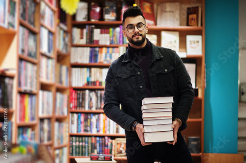 Tall smart arab student man, wear on black jeans jacket and eyeglasses, at library with stack of books.