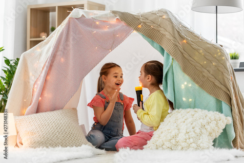 childhood, hygge and friendship concept - happy girls playing with torch light in kids tent at home