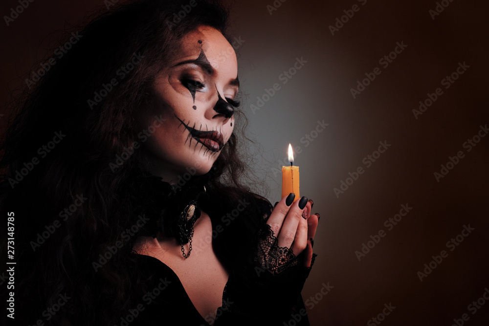 Obraz premium Makeup for Halloween. Portrait of a sexy brunette girl in black clothes with a scary art on her face. Female hands with a burning candle. Pensive look on the flame. Concept in the devilish style.