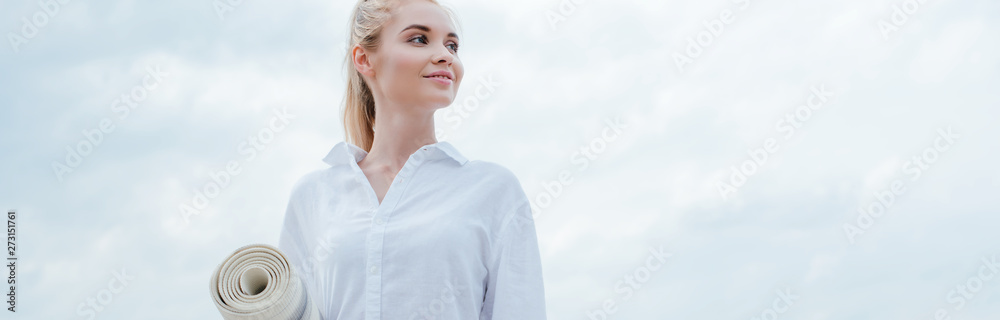 panoramic shot of cheerful young blonde woman standing near sea and holding yoga mat