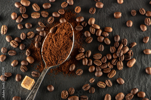 A metal spoon with ground coffee lies on a slate tray, on which coffee seeds are spread out.