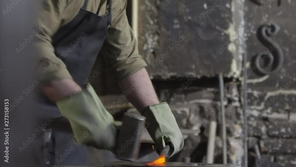 Caucasian craftsman with beard in safety apron and gloves standing at anvil and manually forging molten metal billet with hammer Caucasian craftsman with beard in safety apron and gloves standing at anvil and manually forging molten metal billet with hammer
