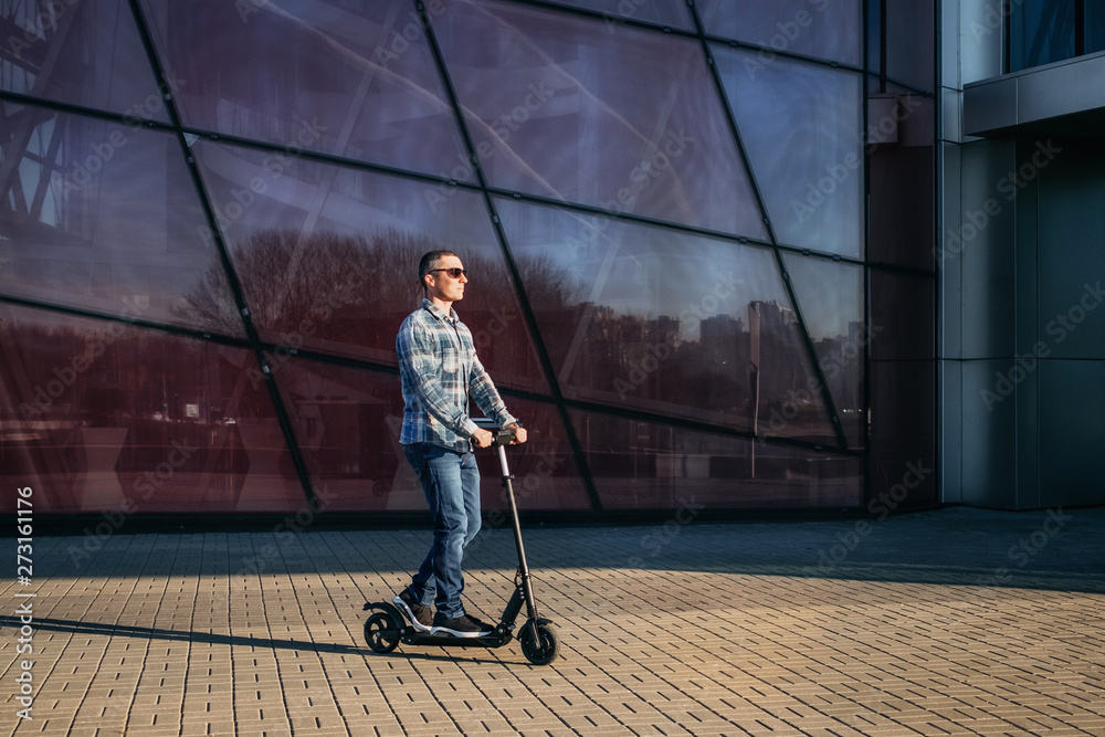 Obraz premium Man riding a electric kick scooter on stone pavement against modern glass wall of building