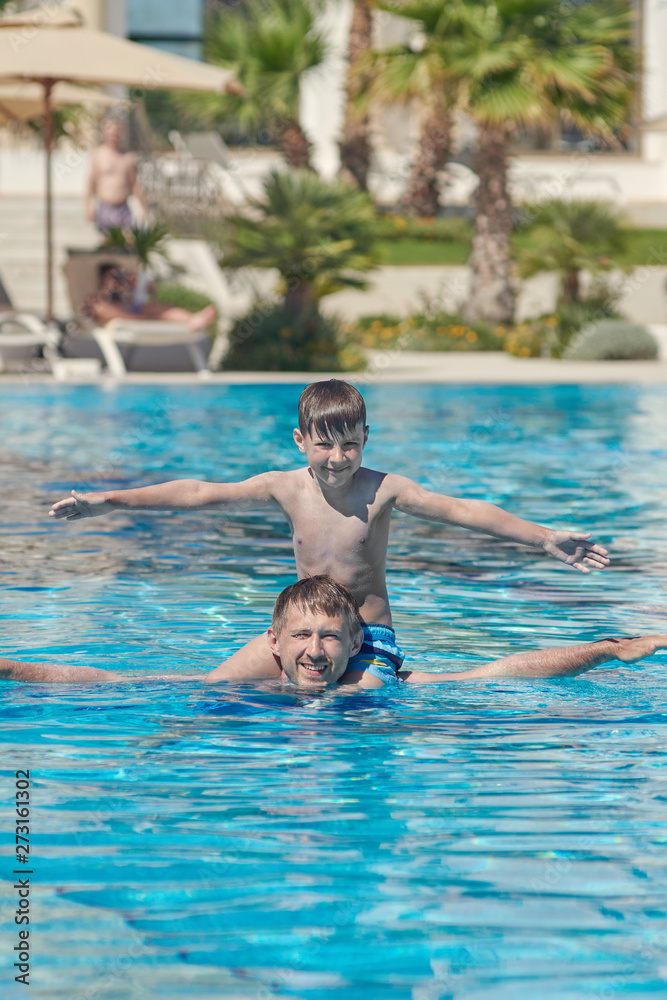 Dad and son having fun in the hotel’s pool. They are smiling and their arms are open wide.