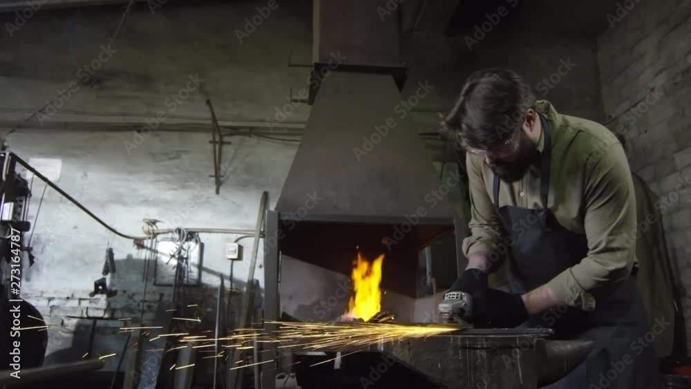 Tracking right of bearded Caucasian blacksmith in safety apron, gloves and glasses standing at anvil and shaping iron detail using hand electrical circular saw with abrasive disk Tracking right of bearded Caucasian blacksmith in safety apron, gloves and glasses standing at anvil and shaping iron detail using hand electrical circular saw with abrasive disk