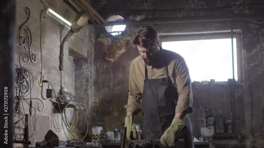 Medium shot of bearded blacksmith standing at anvil, forging metal billet, then taking phone from apron pocket and answering phone call Medium shot of bearded blacksmith standing at anvil, forging metal billet, then taking phone from apron pocket and answering phone call