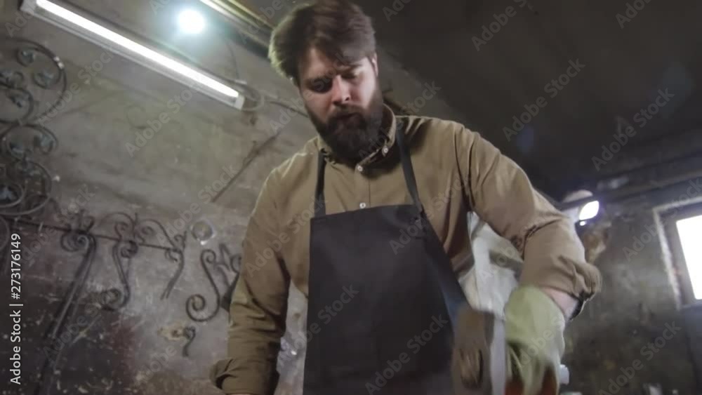 Tilt down of bearded blacksmith wearing apron and gloves standing at anvil and treating hot iron ingot with sledgehammer Tilt down of bearded blacksmith wearing apron and gloves standing at anvil and treating hot iron ingot with sledgehammer