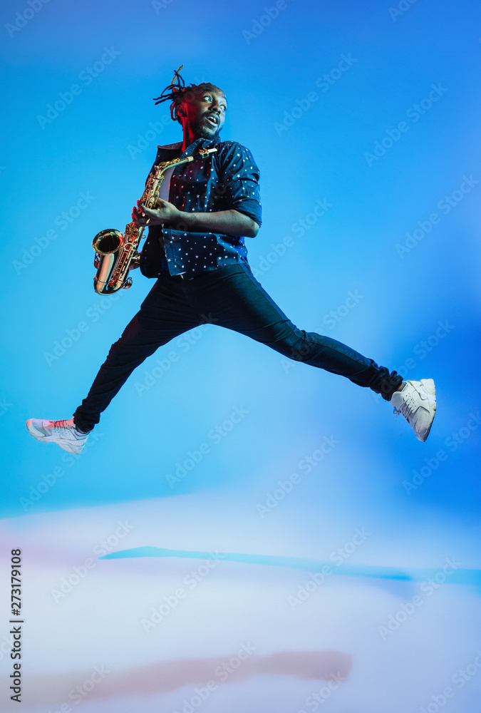 Young african-american jazz musician playing the saxophone on blue studio background in trendy neon light. Concept of music, hobby. Joyful attractive guy improvising. Retro colorful portrait of artist
