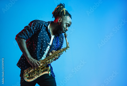 Young african-american jazz musician playing the saxophone on blue studio background in trendy neon light. Concept of music, hobby. Joyful attractive guy improvising. Retro colorful portrait of artist photo