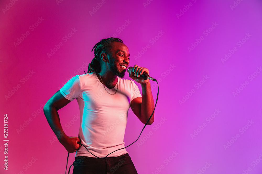 Obraz premium Young african-american jazz musician with microphone singing a song on purple studio background in trendy neon light. Concept of music, hobby, inspirness. Colorful portrait of joyful attractive artist