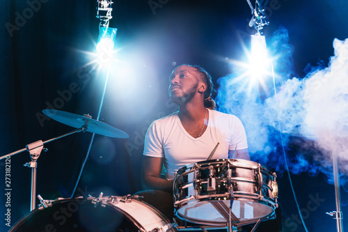 Young african-american jazz musician or drummer playing drums on blue studio background in glowing smoke around him. Concept of music, hobby, inspirness. Colorful portrait of joyful attractive artist.